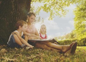 Mother sitting with two children under a tree reading a book
