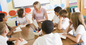 Middle school student sit around a table discussing what they are learning with teacher